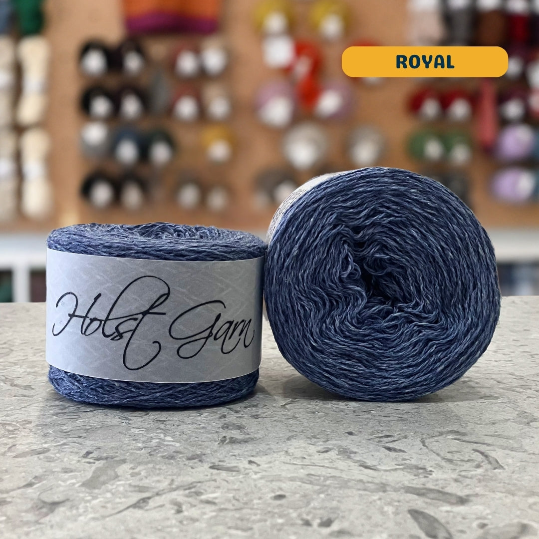 50g royal blue lambswool‑merino‑cotton light fingering skein, 'Holst Garn' label visible, craft shop background (blurred)