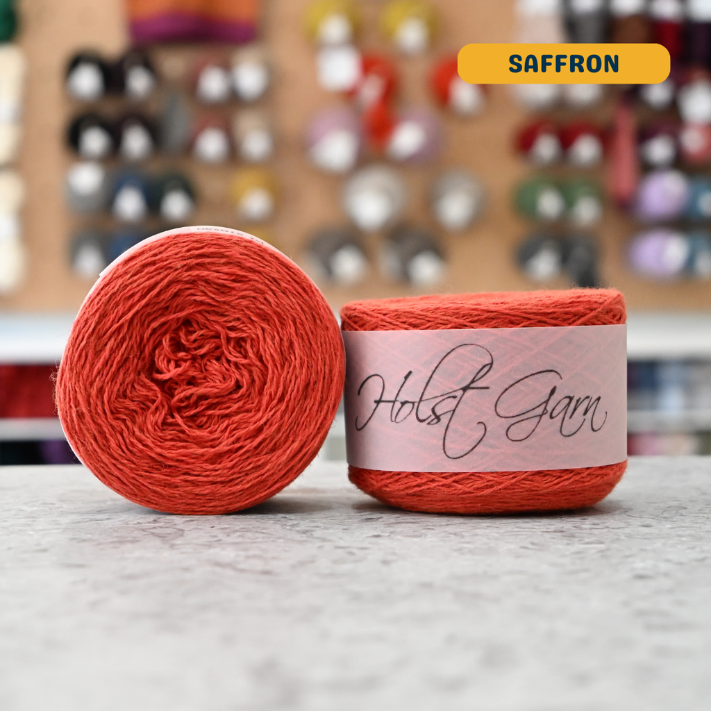 50g orange (saffron) lambswool‑merino‑cotton light fingering skein, 'Holst Garn' label visible, craft shop background (blurred)