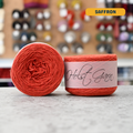 50g orange (saffron) lambswool‑merino‑cotton light fingering skein, 'Holst Garn' label visible, craft shop background (blurred)