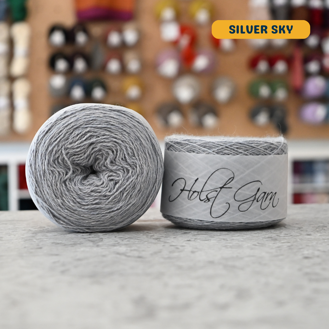 50g light grey (sliver sky) lambswool‑merino‑cotton light fingering skein, 'Holst Garn' label visible, craft shop background (blurred)