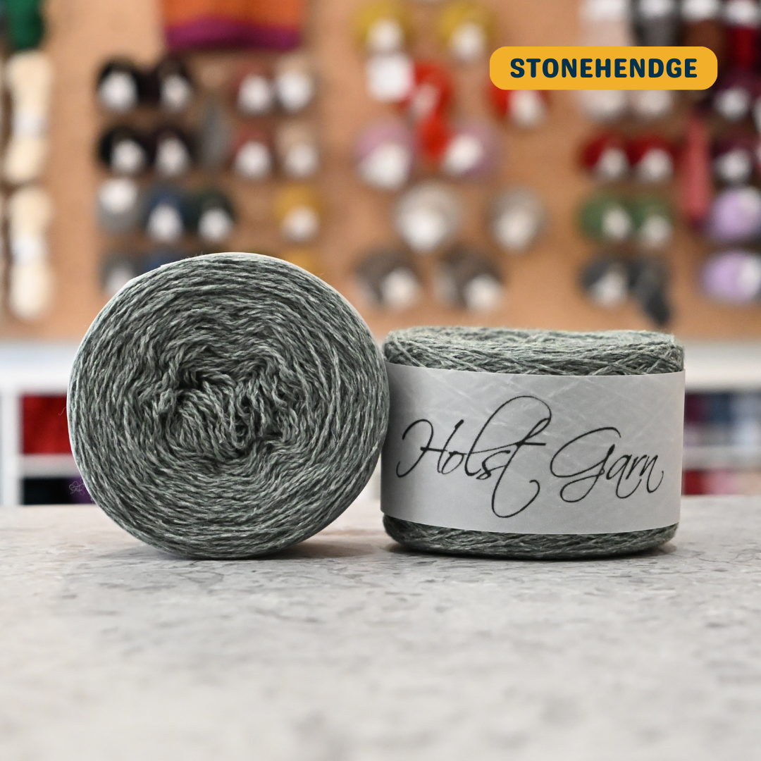 50g sage green (stonehenge) lambswool‑merino‑cotton light fingering skein, 'Holst Garn' label visible, craft shop background (blurred)