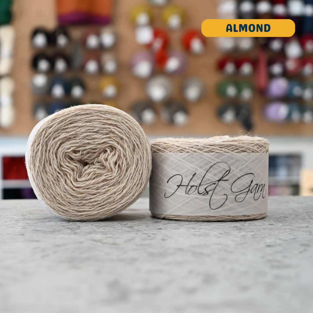 50g light beige (almond) merino & shetland wool light fingering yarn, 'Holst Garn' label visible, craft shop background (blurred)