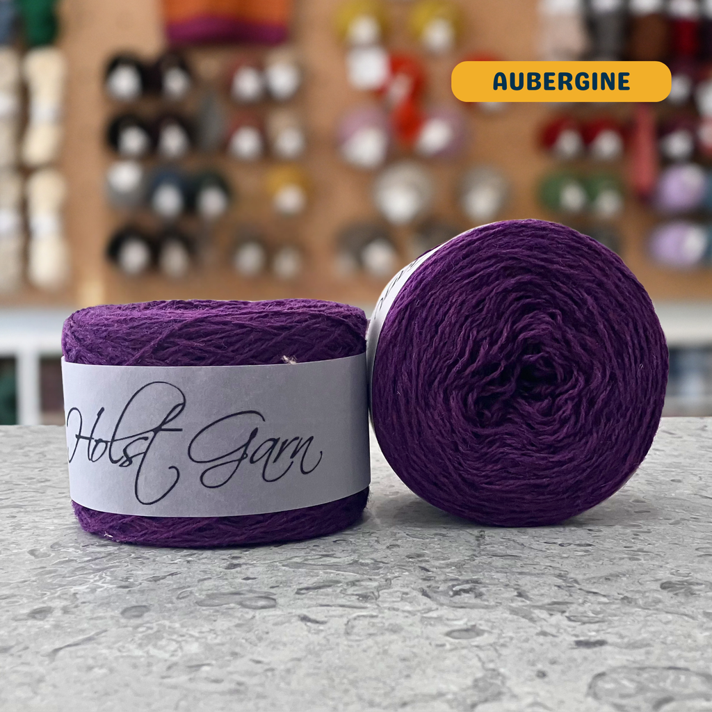 50g dark purple (aubergine) merino & shetland wool light fingering yarn, 'Holst Garn' label visible, craft shop background (blurred)