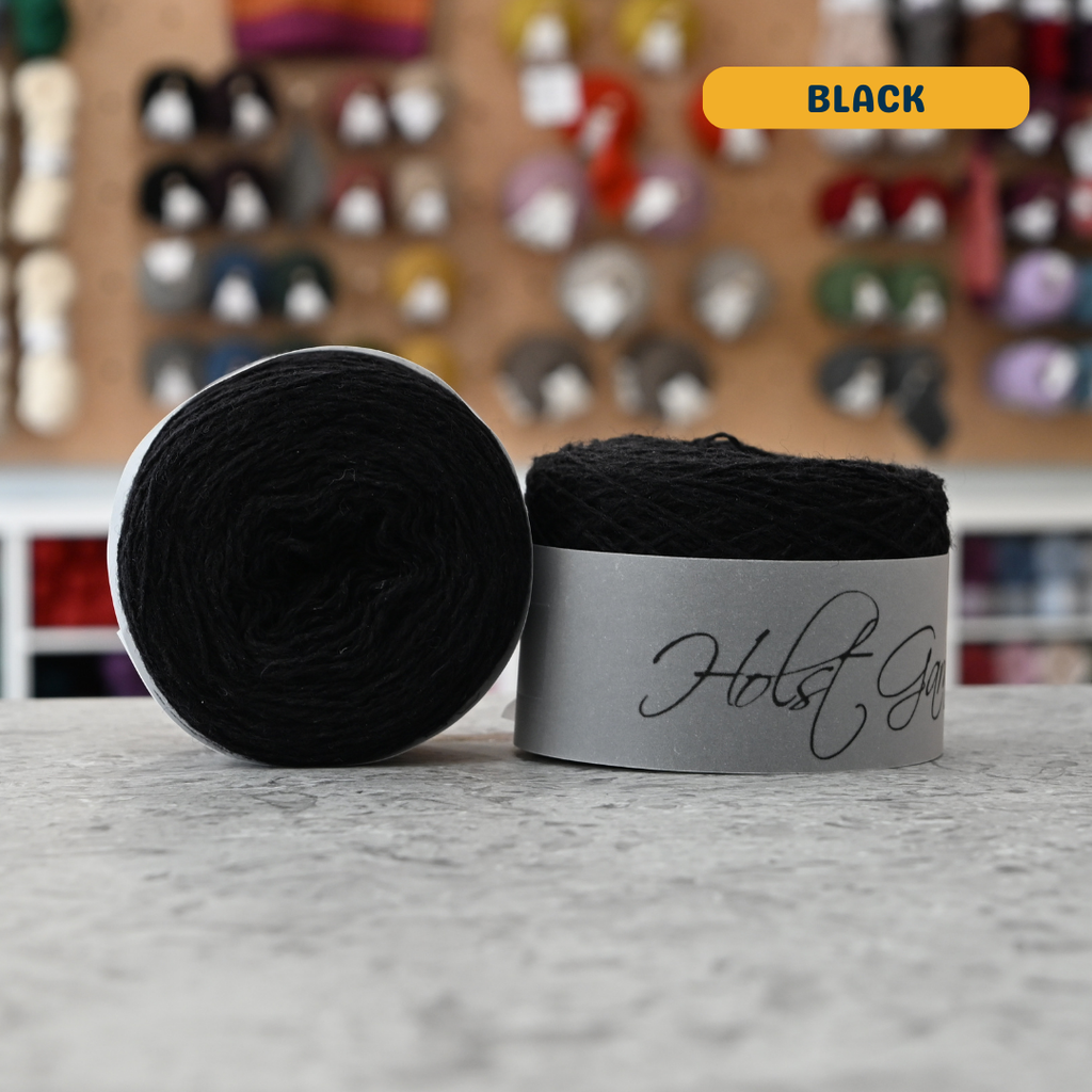 50g black merino & shetland wool light fingering yarn, 'Holst Garn' label visible, craft shop background (blurred)