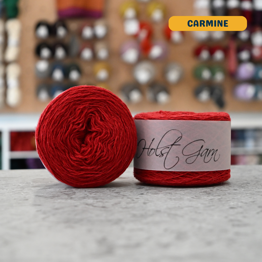 50g red (carmine) merino & shetland wool light fingering yarn, 'Holst Garn' label visible, craft shop background (blurred)