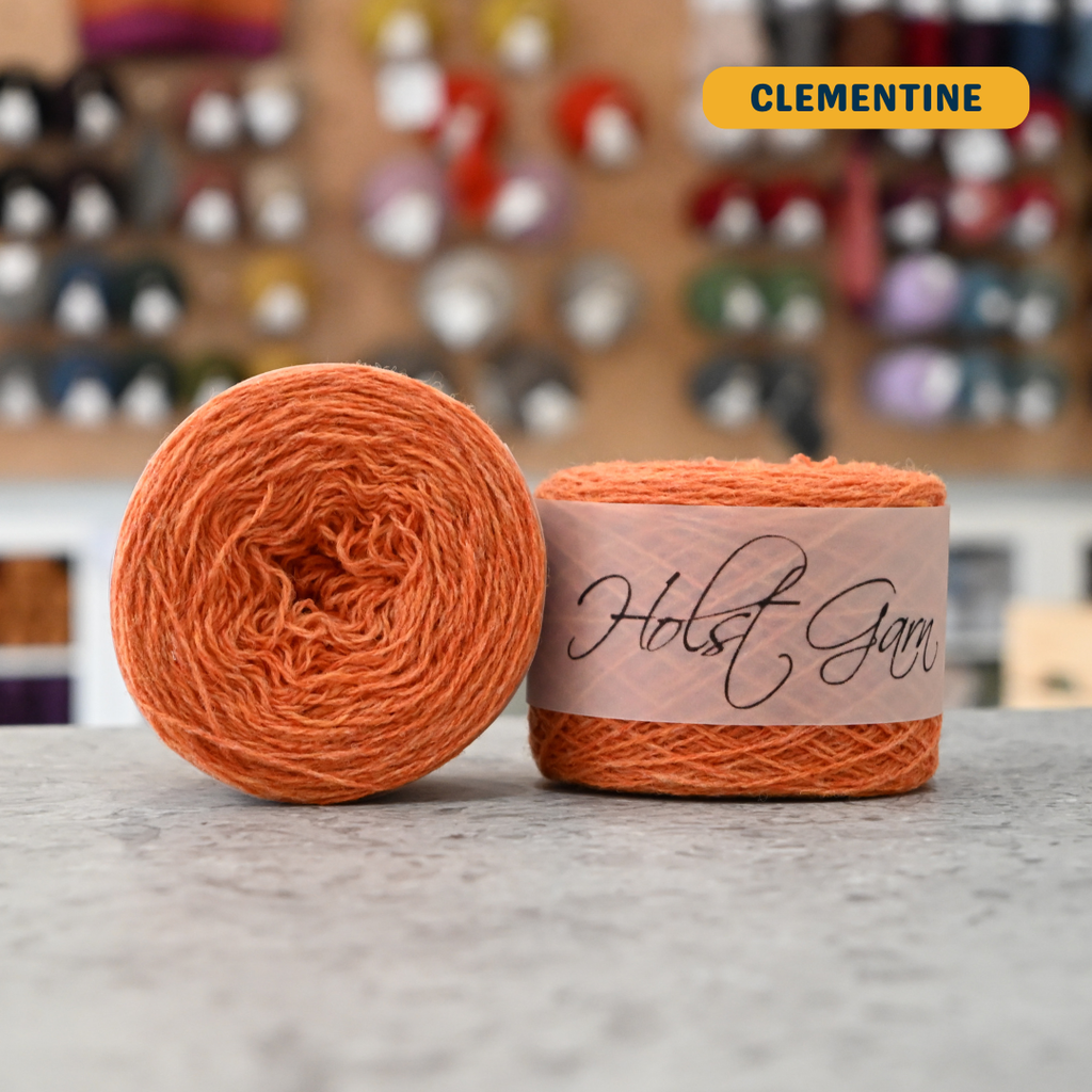 50g bright orange (clementine) merino & shetland wool light fingering yarn, 'Holst Garn' label visible, craft shop background (blurred)