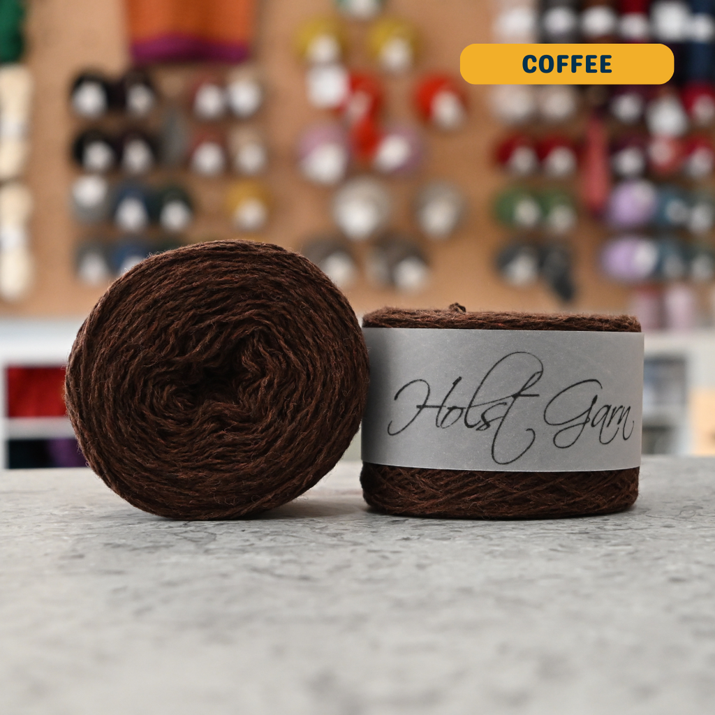 50g brown (coffee) merino & shetland wool light fingering yarn, 'Holst Garn' label visible, craft shop background (blurred)