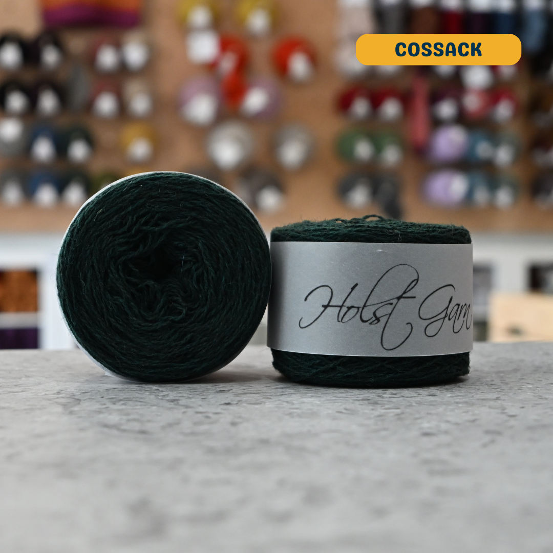 50g dark green (cossack) merino & shetland wool light fingering yarn, 'Holst Garn' label visible, craft shop background (blurred)