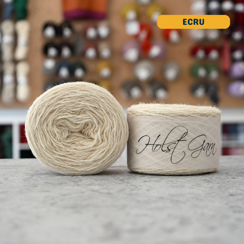 50g natural white (ecru) merino & shetland wool light fingering yarn, 'Holst Garn' label visible, craft shop background (blurred)