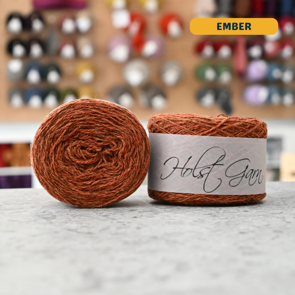 50g burnt orange (ember) merino & shetland wool light fingering yarn, 'Holst Garn' label visible, craft shop background (blurred)