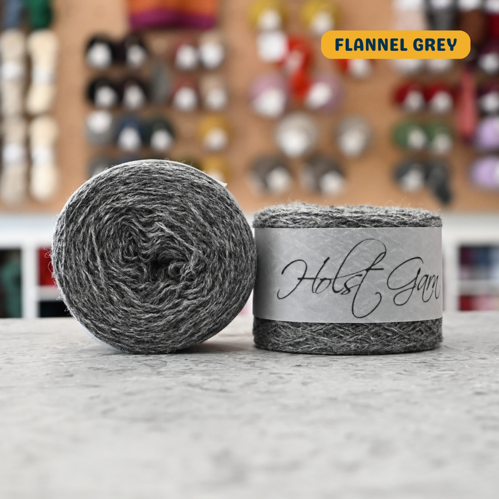 50g medium grey (flannel grey) merino & shetland wool light fingering yarn, 'Holst Garn' label visible, craft shop background (blurred)