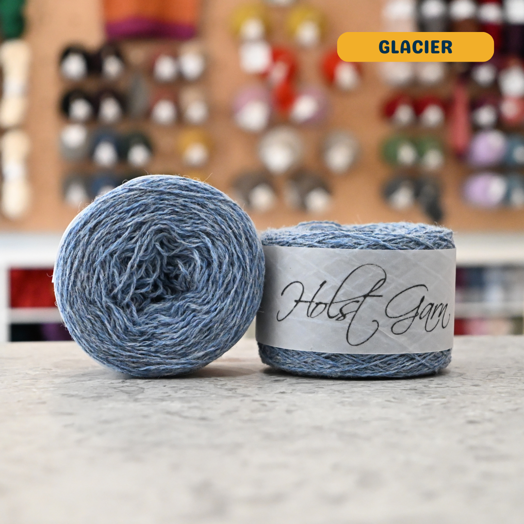50g light blue (glacier) merino & shetland wool light fingering yarn, 'Holst Garn' label visible, craft shop background (blurred)