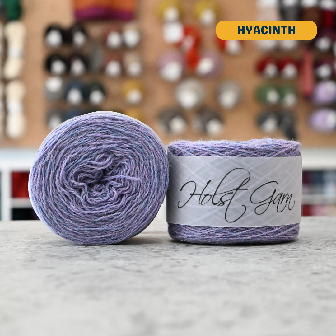 50g light blue-purple (hyacinth) merino & shetland wool light fingering yarn, 'Holst Garn' label visible, craft shop background (blurred)