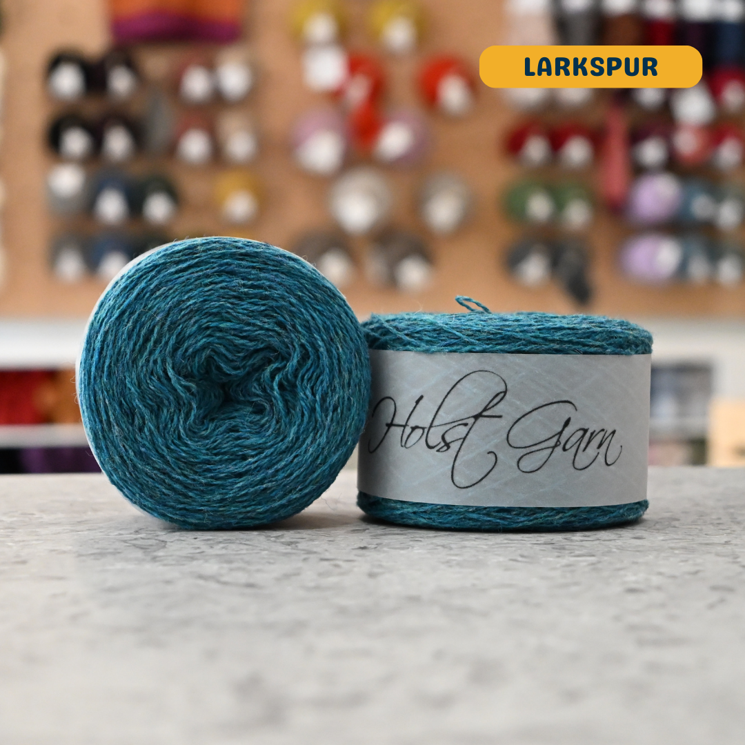50g vibrant blue (larkspur) merino & shetland wool light fingering yarn, 'Holst Garn' label visible, craft shop background (blurred)