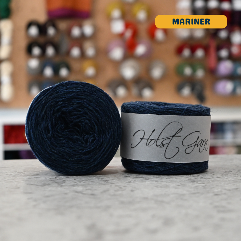 50g navy blue (mariner) merino & shetland wool light fingering yarn, 'Holst Garn' label visible, craft shop background (blurred)