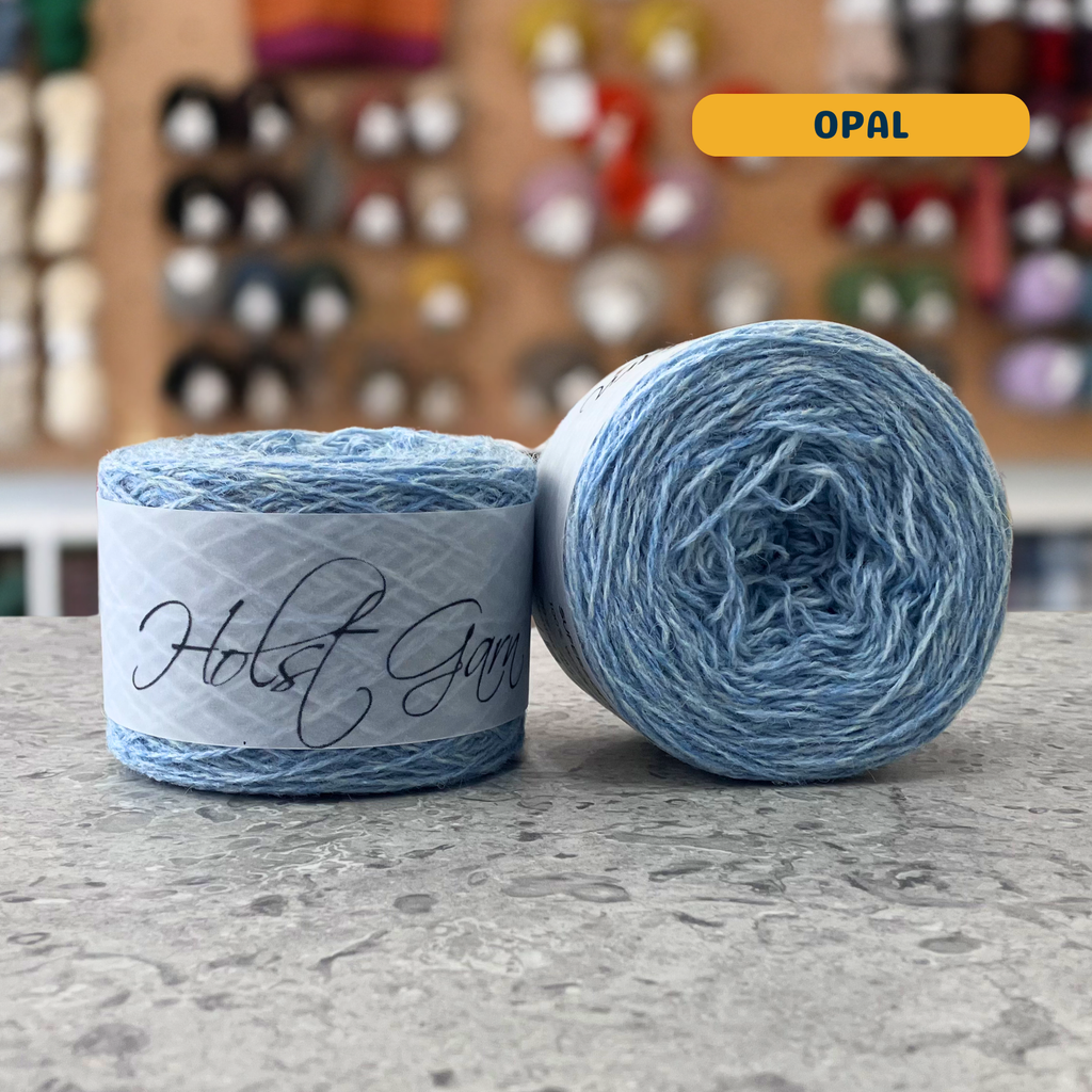 50g light blue (opal) merino & shetland wool light fingering yarn, 'Holst Garn' label visible, craft shop background (blurred)