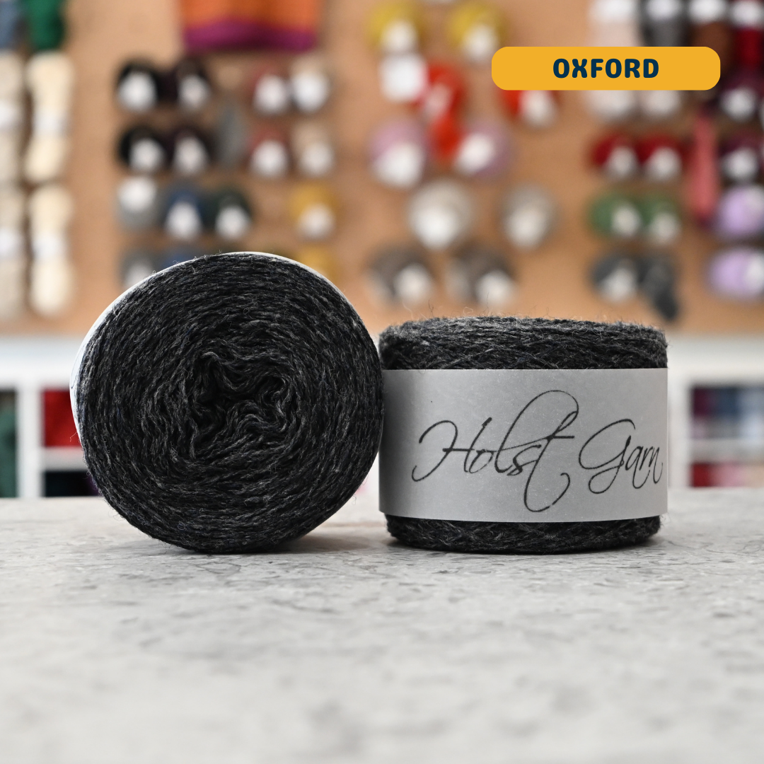 50g dark grey (oxford) merino & shetland wool light fingering yarn, 'Holst Garn' label visible, craft shop background (blurred)