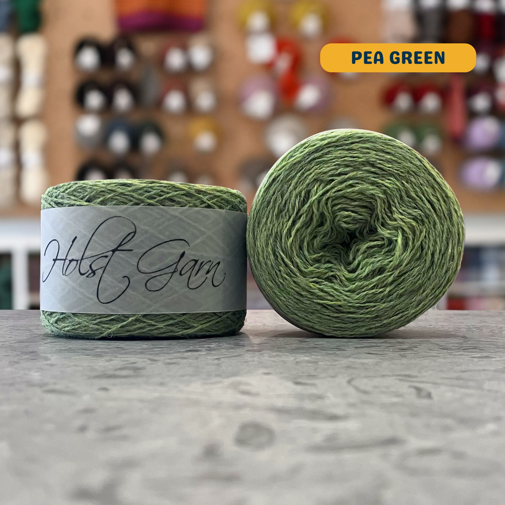 50g green (pea green) merino & shetland wool light fingering yarn, 'Holst Garn' label visible, craft shop background (blurred)