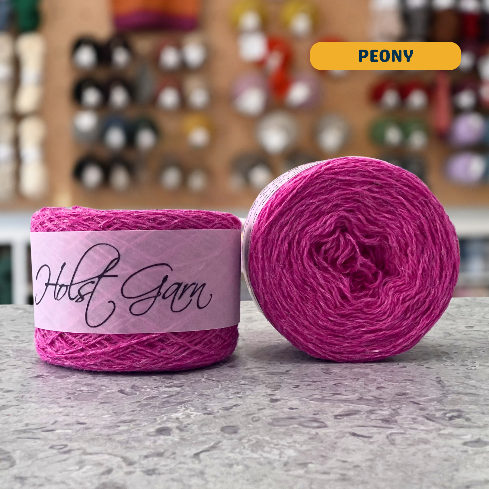 50g vibrant pink (peony) merino & shetland wool light fingering yarn, 'Holst Garn' label visible, craft shop background (blurred)