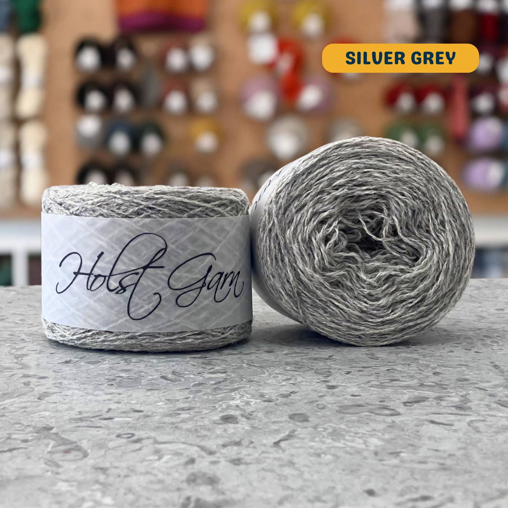 50g light grey (silver grey) merino & shetland wool light fingering yarn, 'Holst Garn' label visible, craft shop background (blurred)