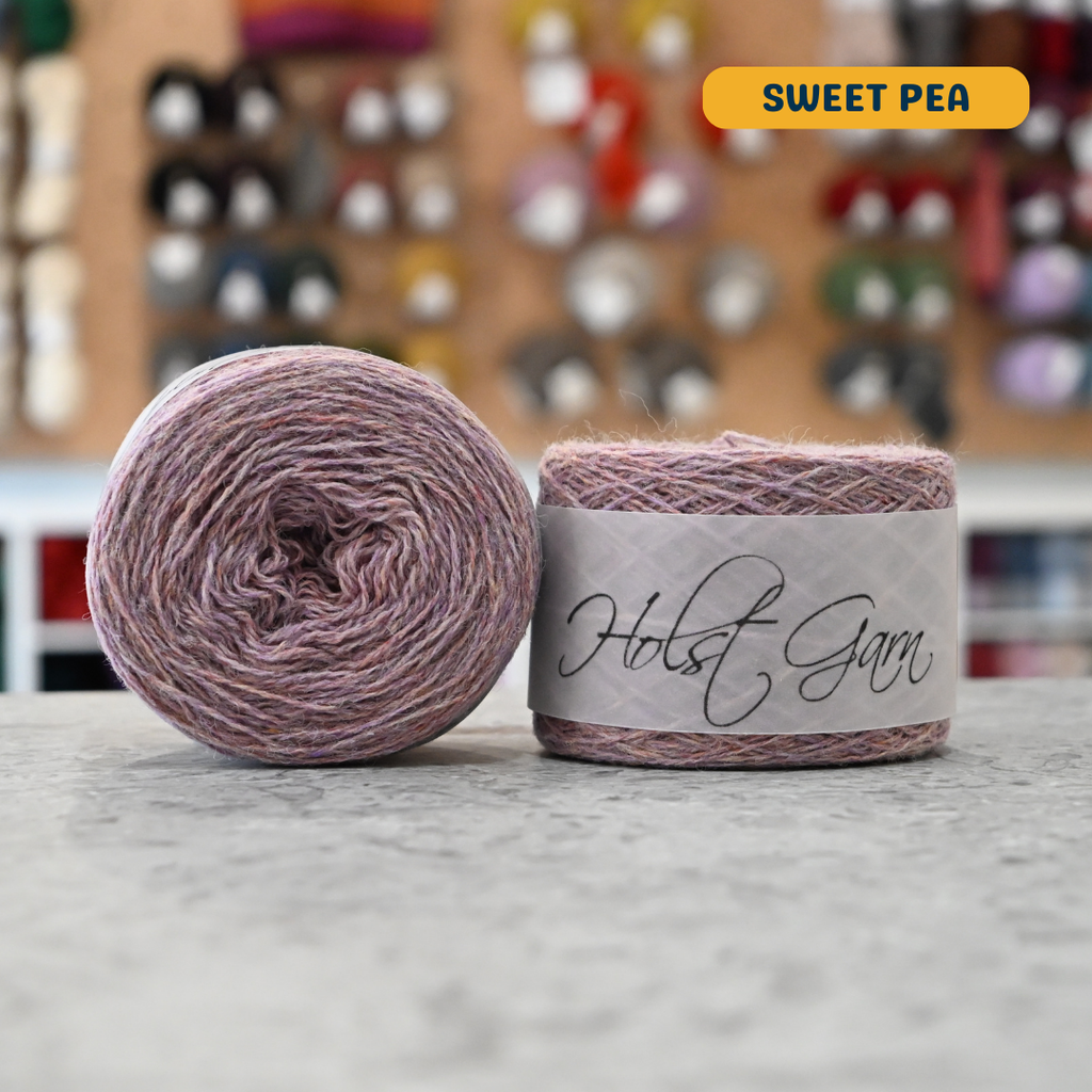 50g light grey-pink (sweet pea) merino & shetland wool light fingering yarn, 'Holst Garn' label visible, craft shop background (blurred)