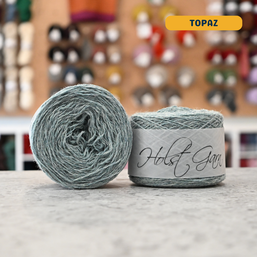 50g light green-blue (topaz) merino & shetland wool light fingering yarn, 'Holst Garn' label visible, craft shop background (blurred)