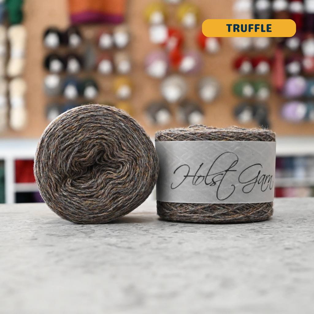50g brown-grey (truffle) merino & shetland wool light fingering yarn, 'Holst Garn' label visible, craft shop background (blurred)