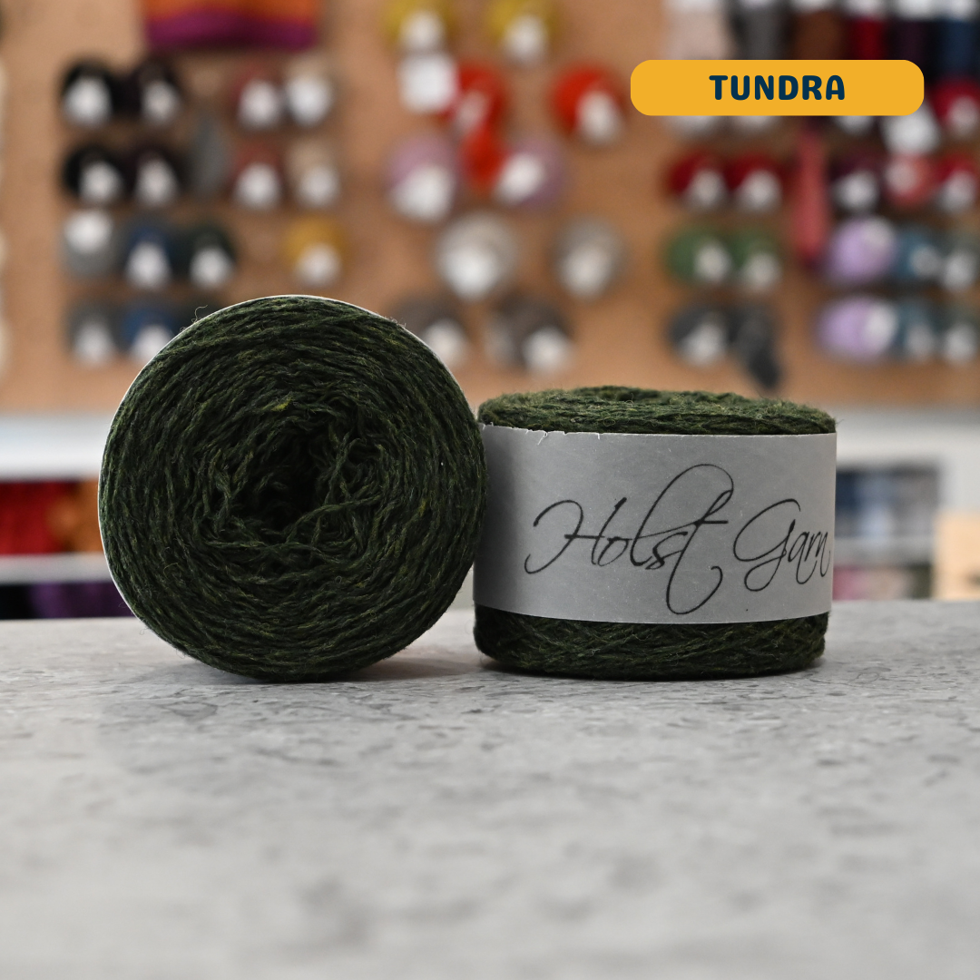 50g dark green (tundra) merino & shetland wool light fingering yarn, 'Holst Garn' label visible, craft shop background (blurred)