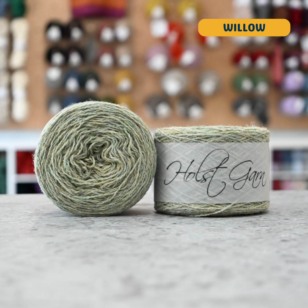 50g light green (willow) merino & shetland wool light fingering yarn, 'Holst Garn' label visible, craft shop background (blurred)