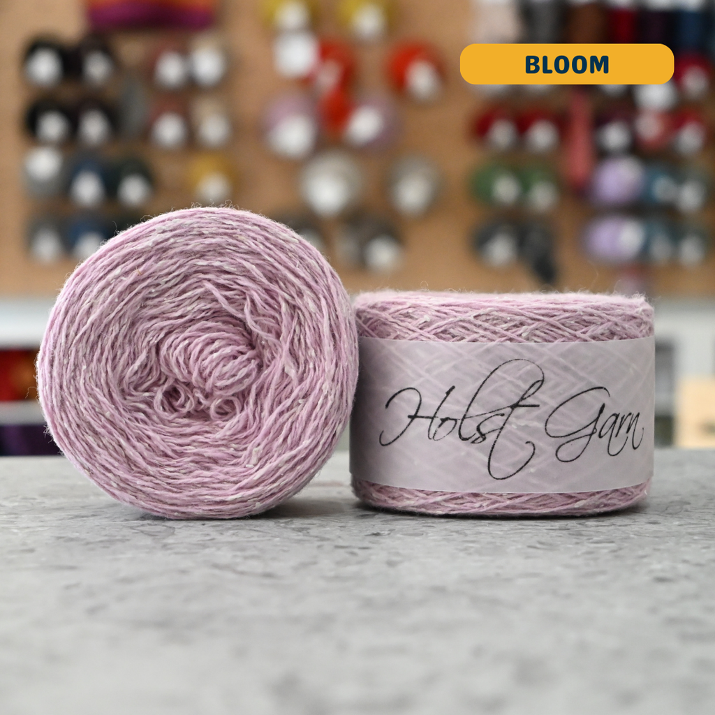 50g light pink (bloom) merino‑silk light fingering skein, 'Holst Garn' label visible, craft shop background (blurred)
