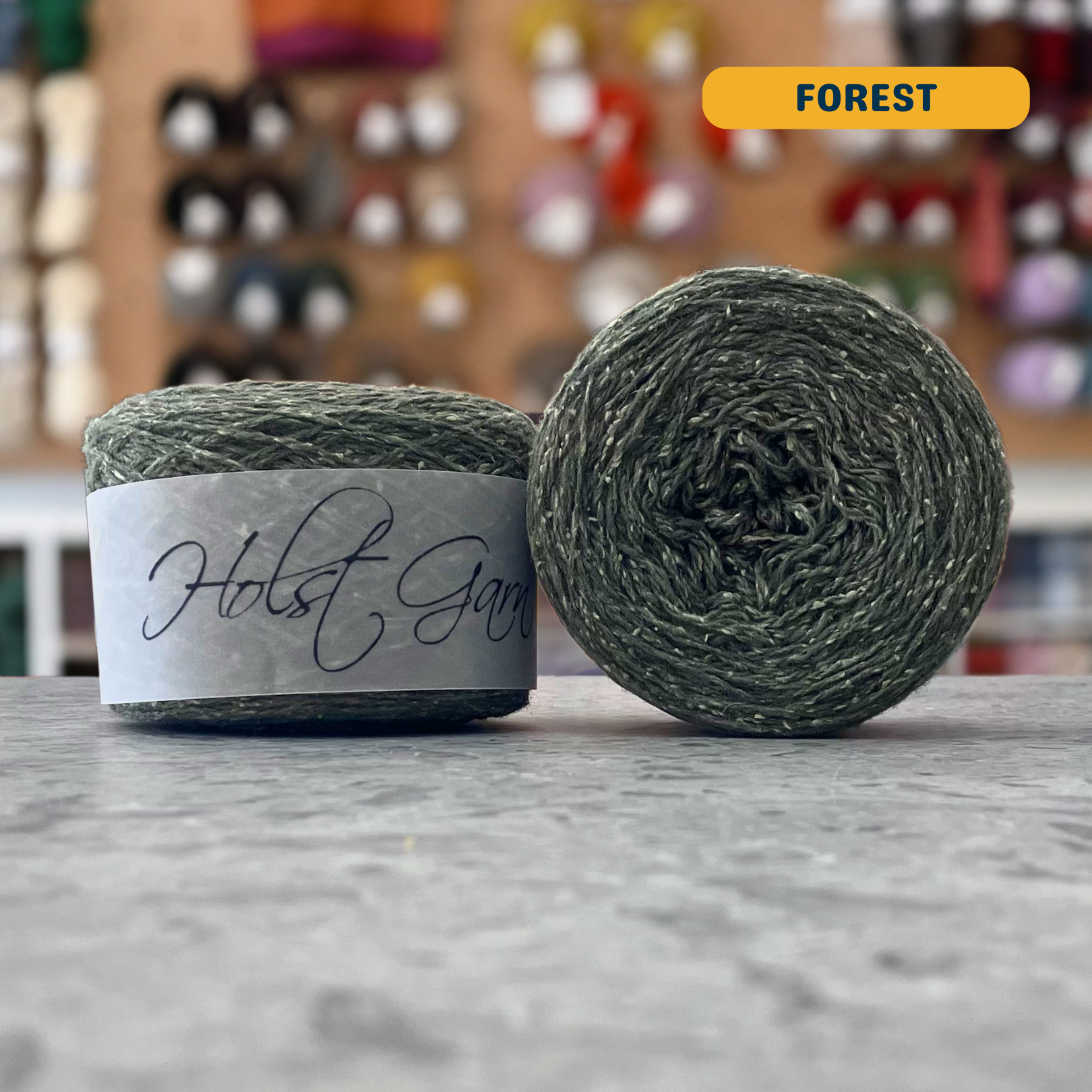 50g green (forest) merino‑silk light fingering skein, 'Holst Garn' label visible, craft shop background (blurred)