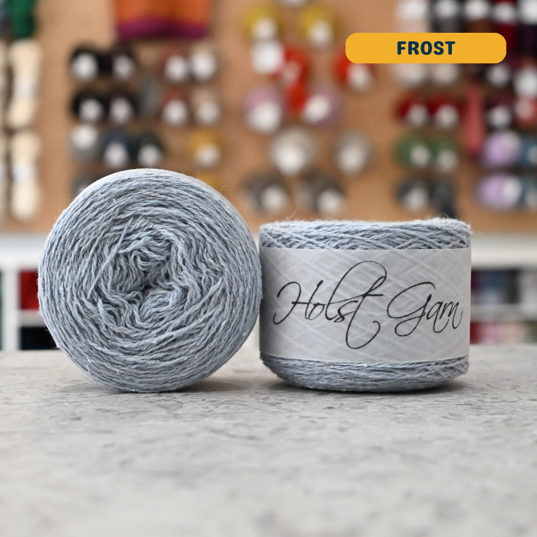 50g light blue (frost) merino‑silk light fingering skein, 'Holst Garn' label visible, craft shop background (blurred)
