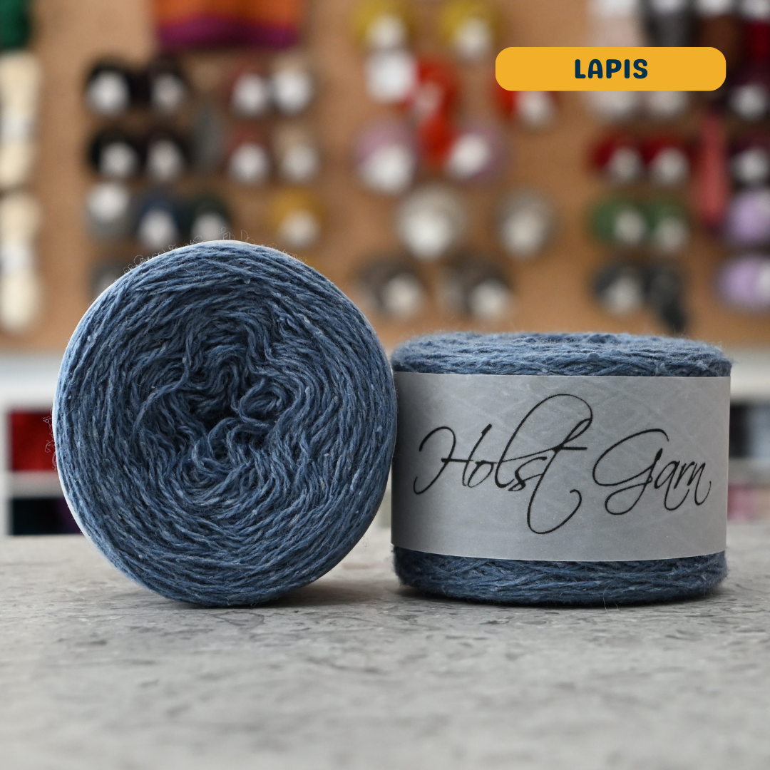 50g blue (lapis) merino‑silk light fingering skein, 'Holst Garn' label visible, craft shop background (blurred)