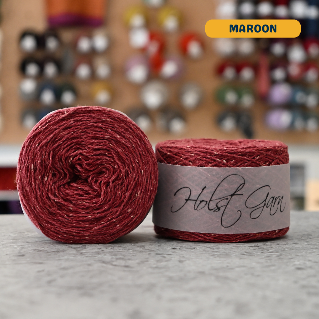 50g red (maroon) merino‑silk tweed light fingering skein, 'Holst Garn' label visible, craft shop background (blurred)