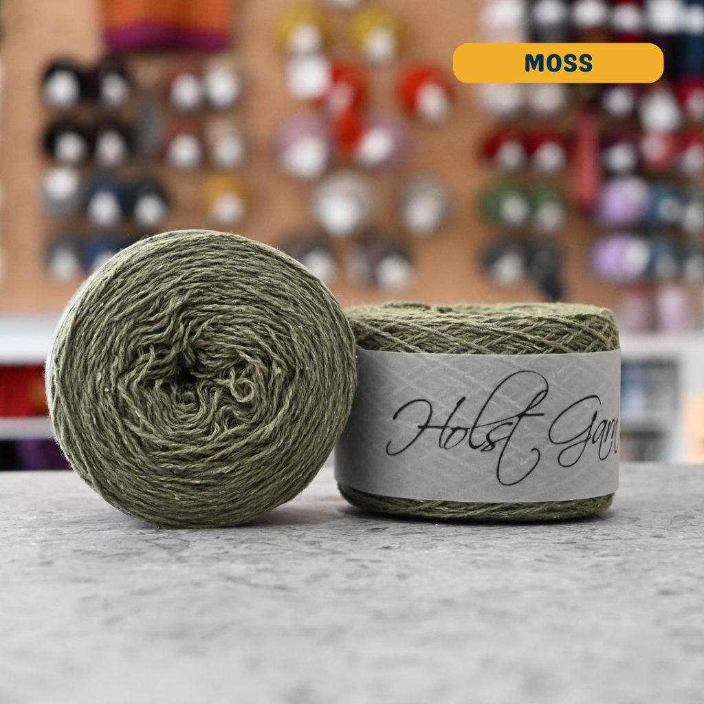 50g sage green (moss) merino‑silk light fingering skein, 'Holst Garn' label visible, craft shop background (blurred)