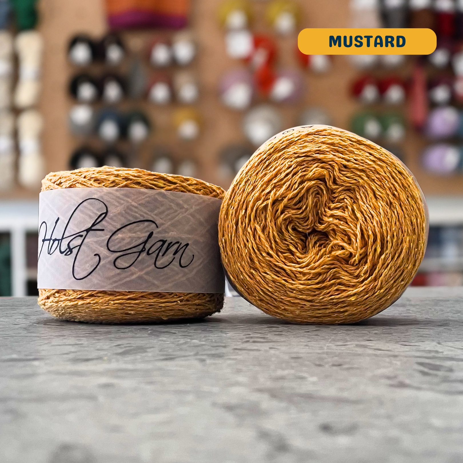 50g mustard yellow merino‑silk light fingering skein, 'Holst Garn' label visible, craft shop background (blurred)