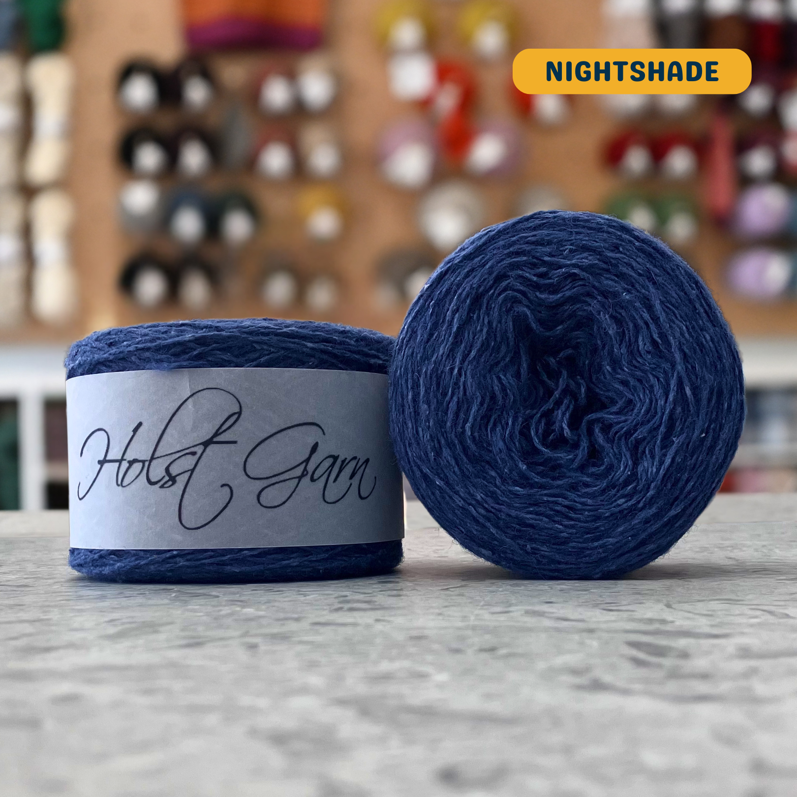 50g dark blue (nightshade) merino‑silk light fingering skein, 'Holst Garn' label visible, craft shop background (blurred)