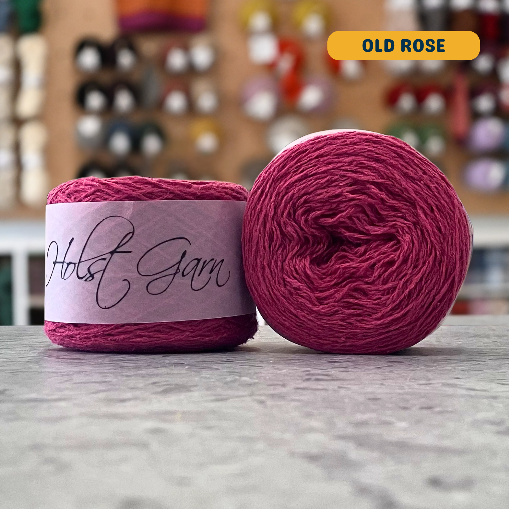 50g pink (old rose) merino‑silk light fingering skein, 'Holst Garn' label visible, craft shop background (blurred)