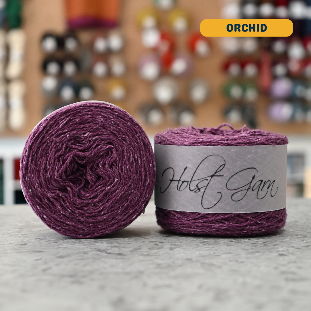 50g purple (orchid) merino‑silk light fingering skein, 'Holst Garn' label visible, craft shop background (blurred)