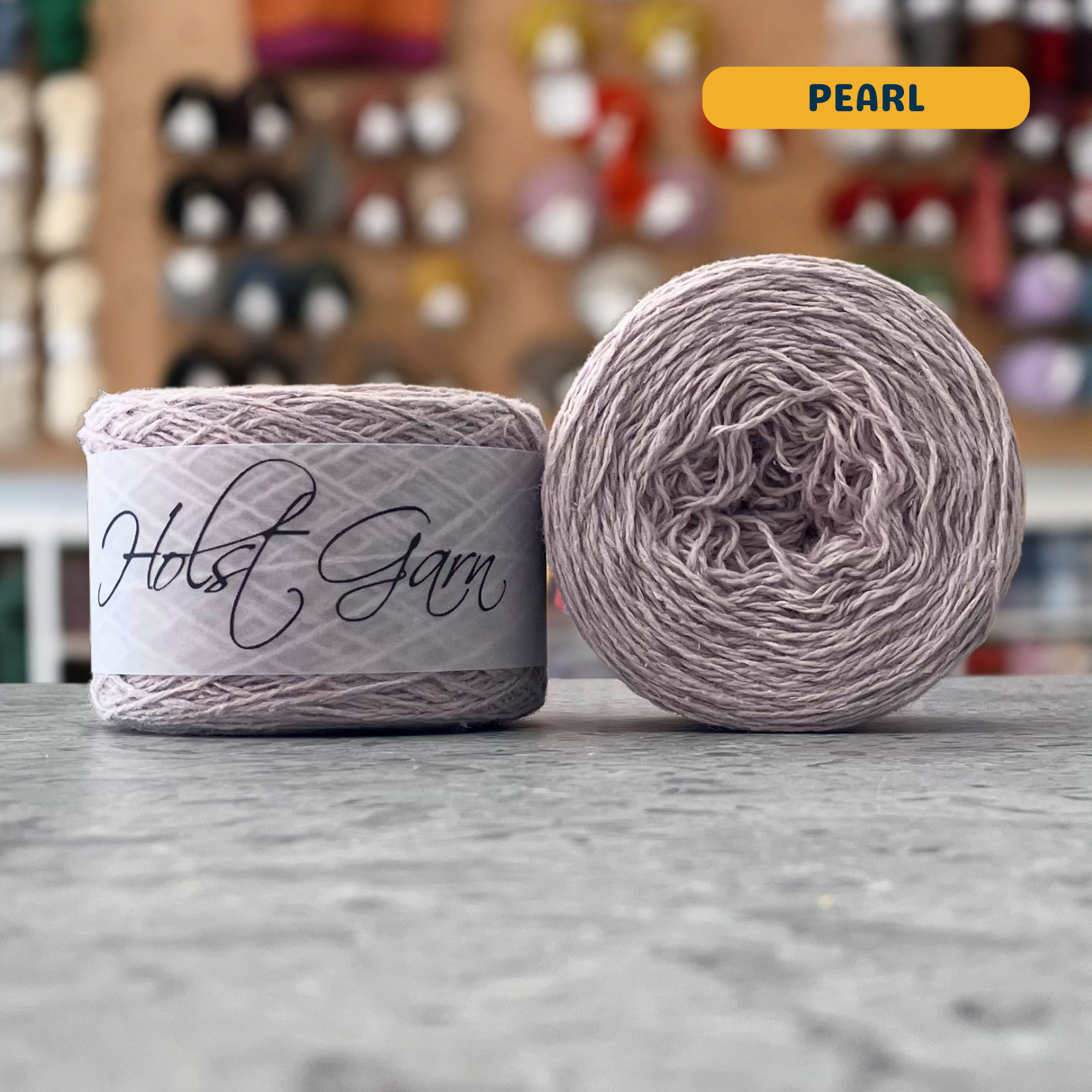 50g cream (pearl) merino‑silk light fingering skein, 'Holst Garn' label visible, craft shop background (blurred)