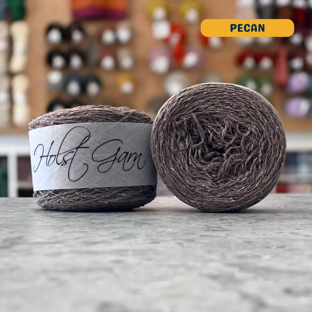 50g light brown (pecan) merino‑silk light fingering skein, 'Holst Garn' label visible, craft shop background (blurred)
