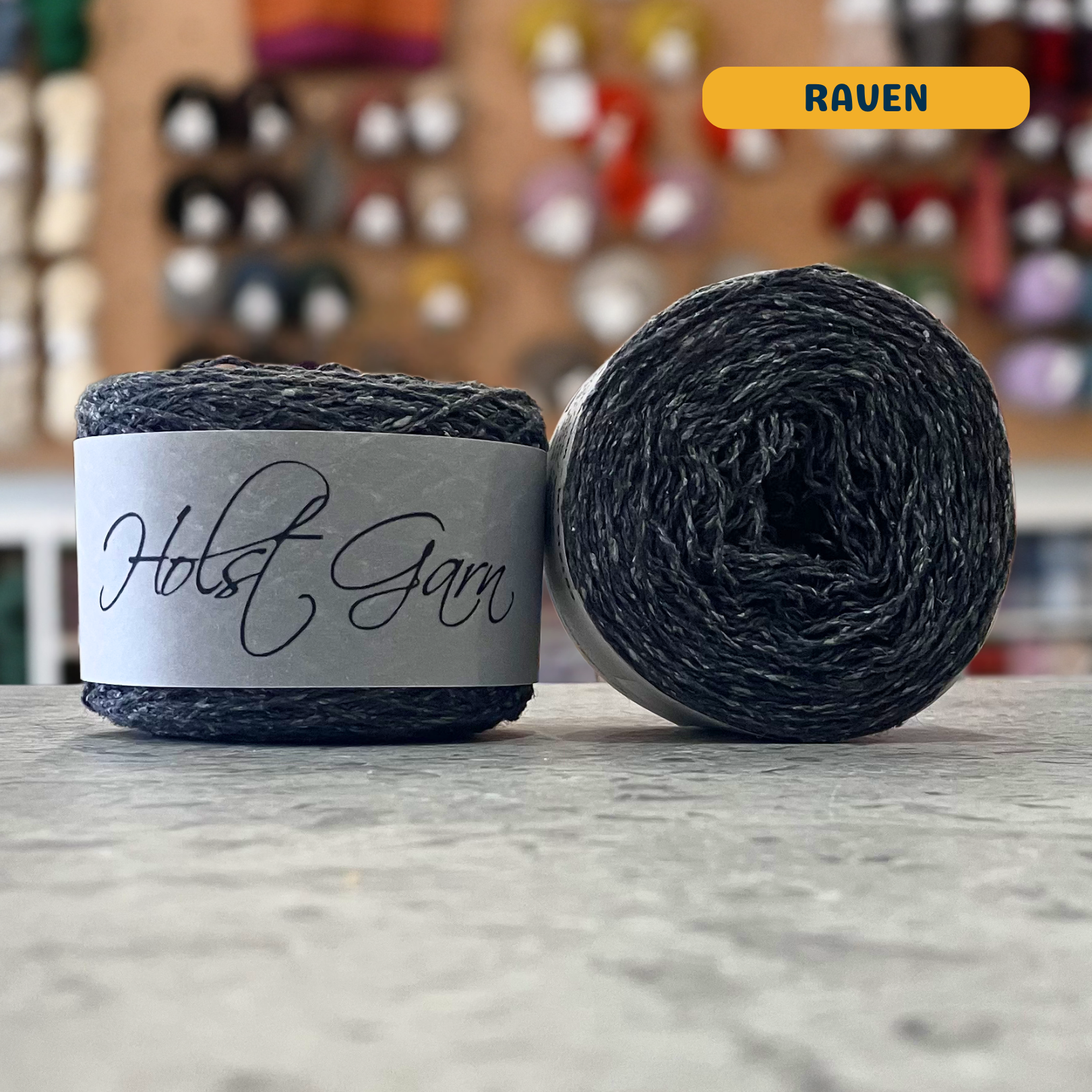50g dark grey (raven) merino‑silk light fingering skein, 'Holst Garn' label visible, craft shop background (blurred)