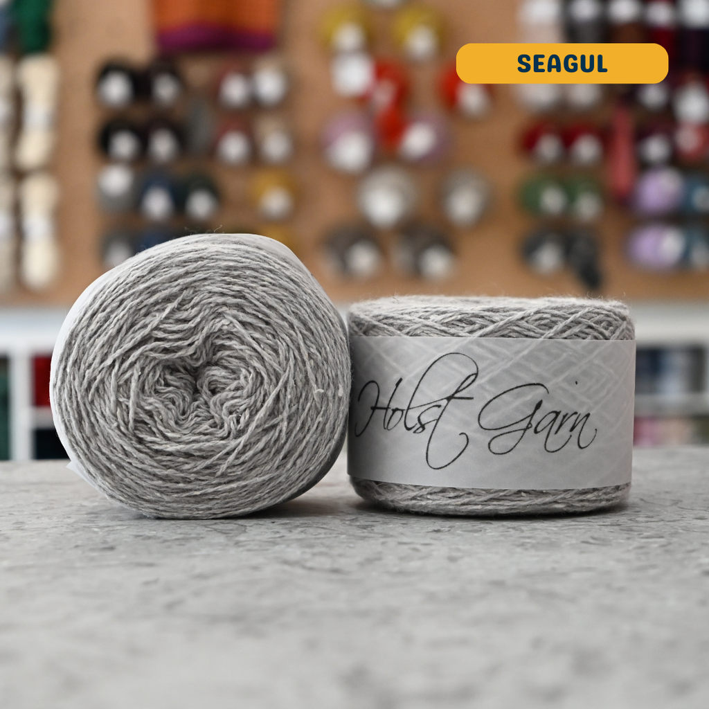 50g light grey (seagul) merino‑silk light fingering skein, 'Holst Garn' label visible, craft shop background (blurred)