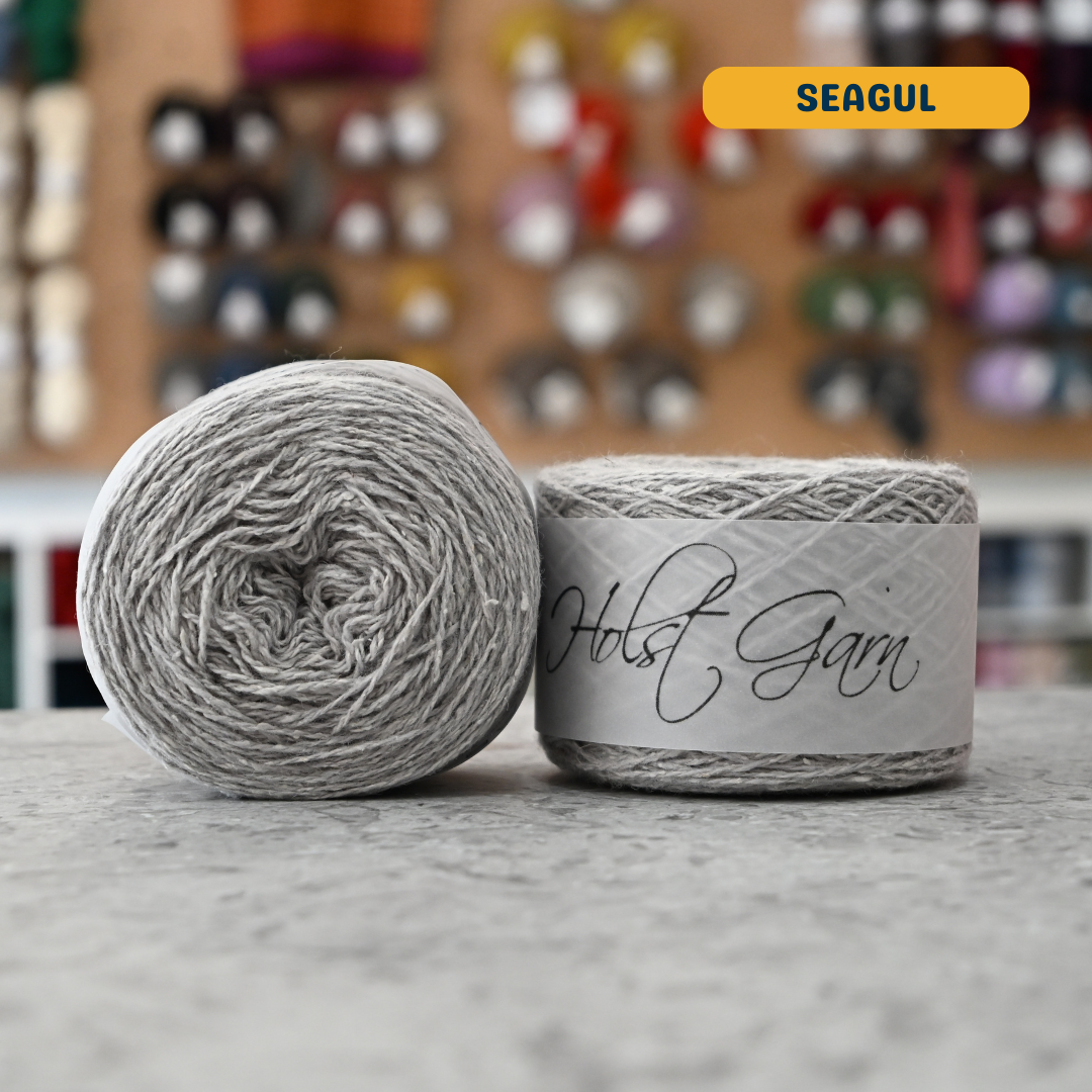 50g light grey (seagul) merino‑silk light fingering skein, 'Holst Garn' label visible, craft shop background (blurred)