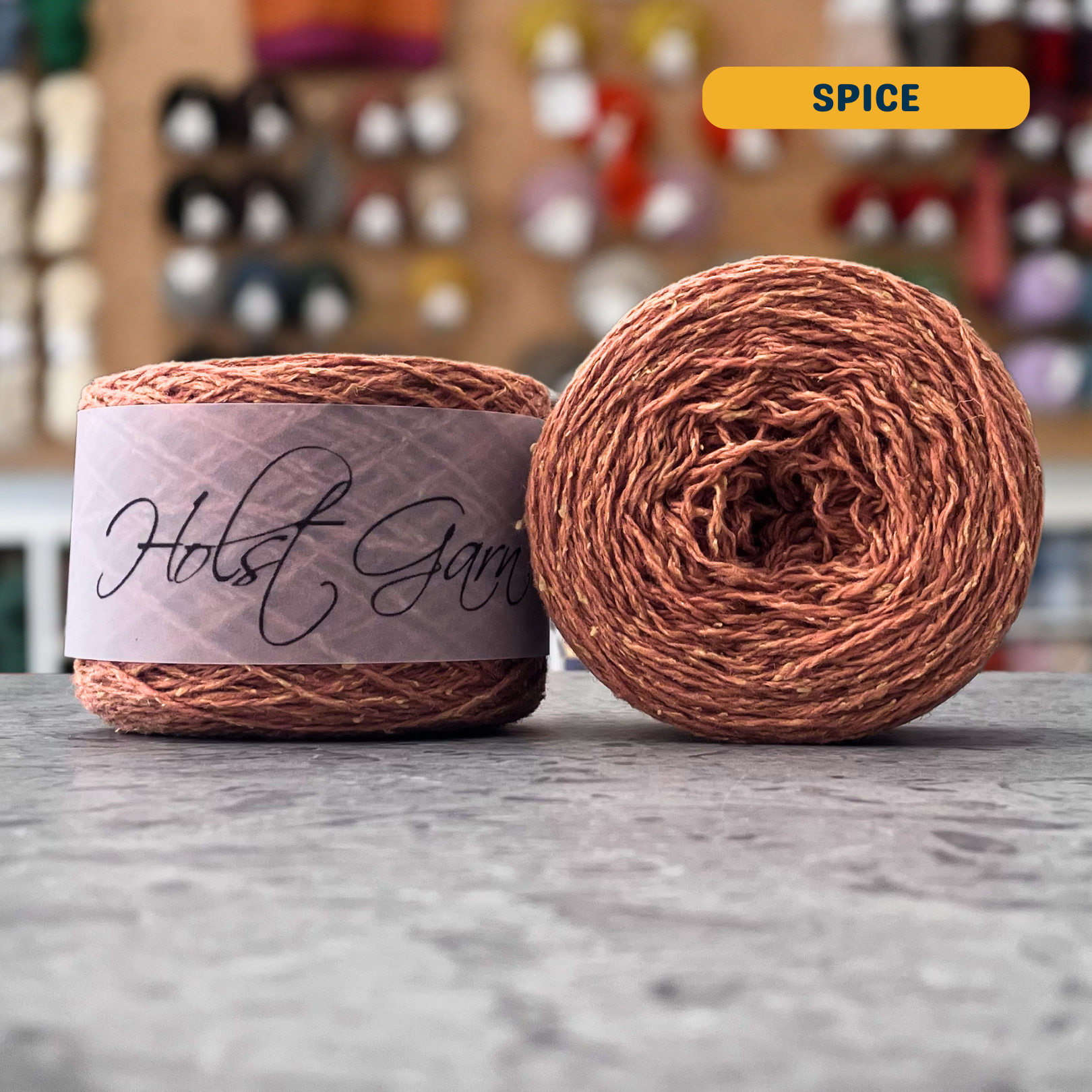 50g orange (spice) merino‑silk light fingering skein, 'Holst Garn' label visible, craft shop background (blurred)
