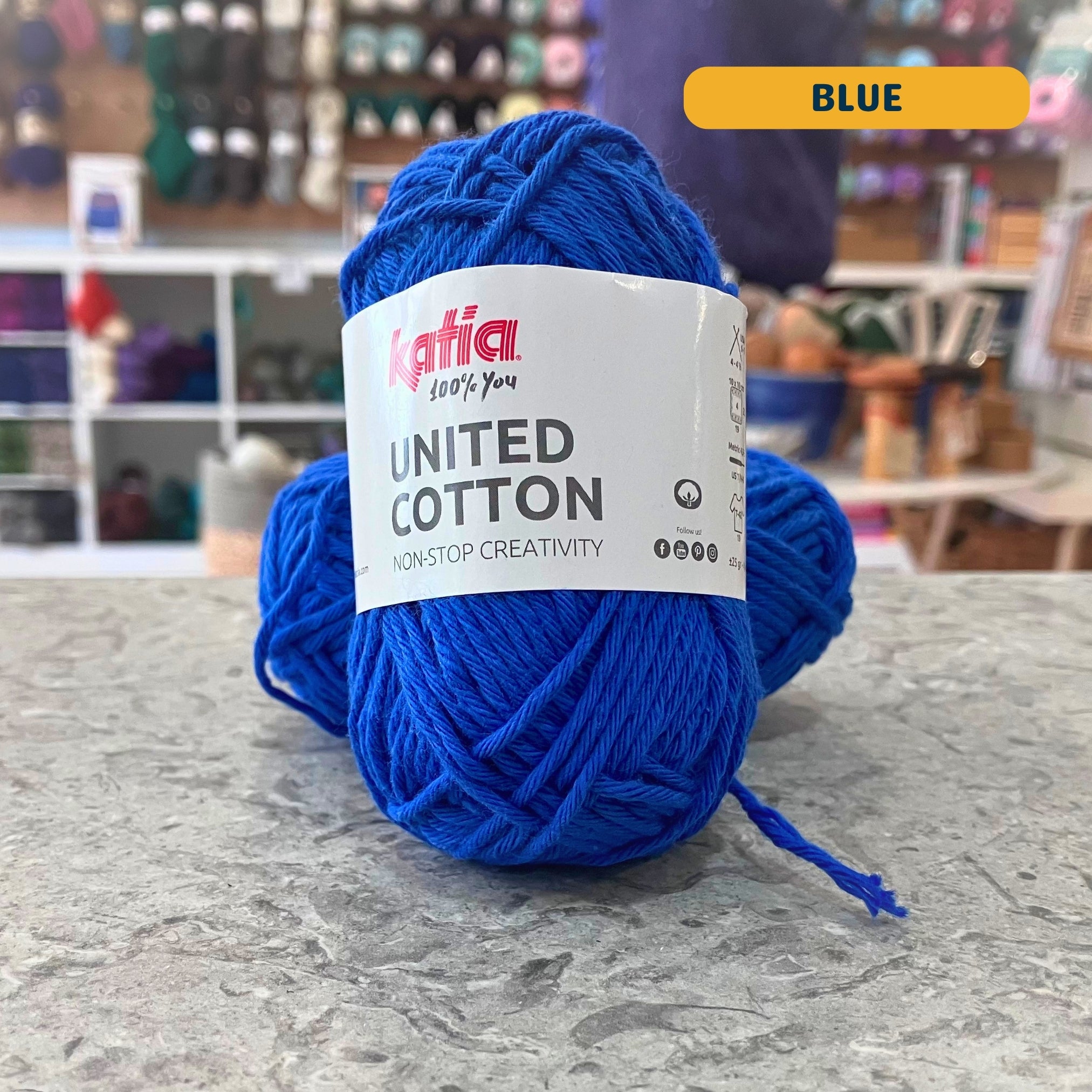 25g royal (blue) cotton DK yarn, 'Katia United Cotton' label visible, craft shop background (blurred)