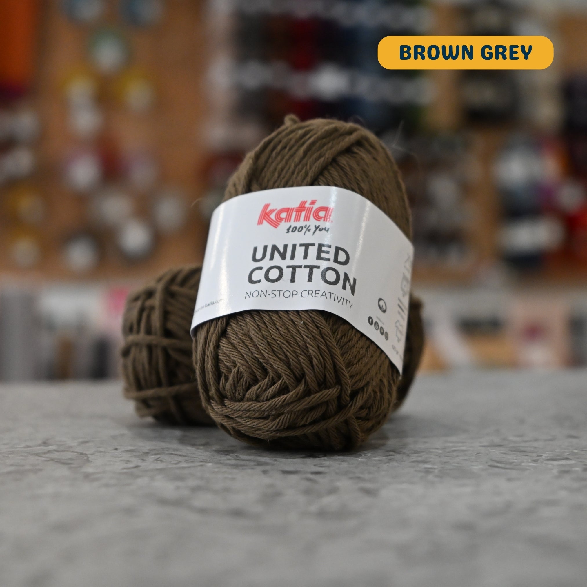 25g dark olive (brown grey) cotton DK yarn, 'Katia United Cotton' label visible, craft shop background (blurred)