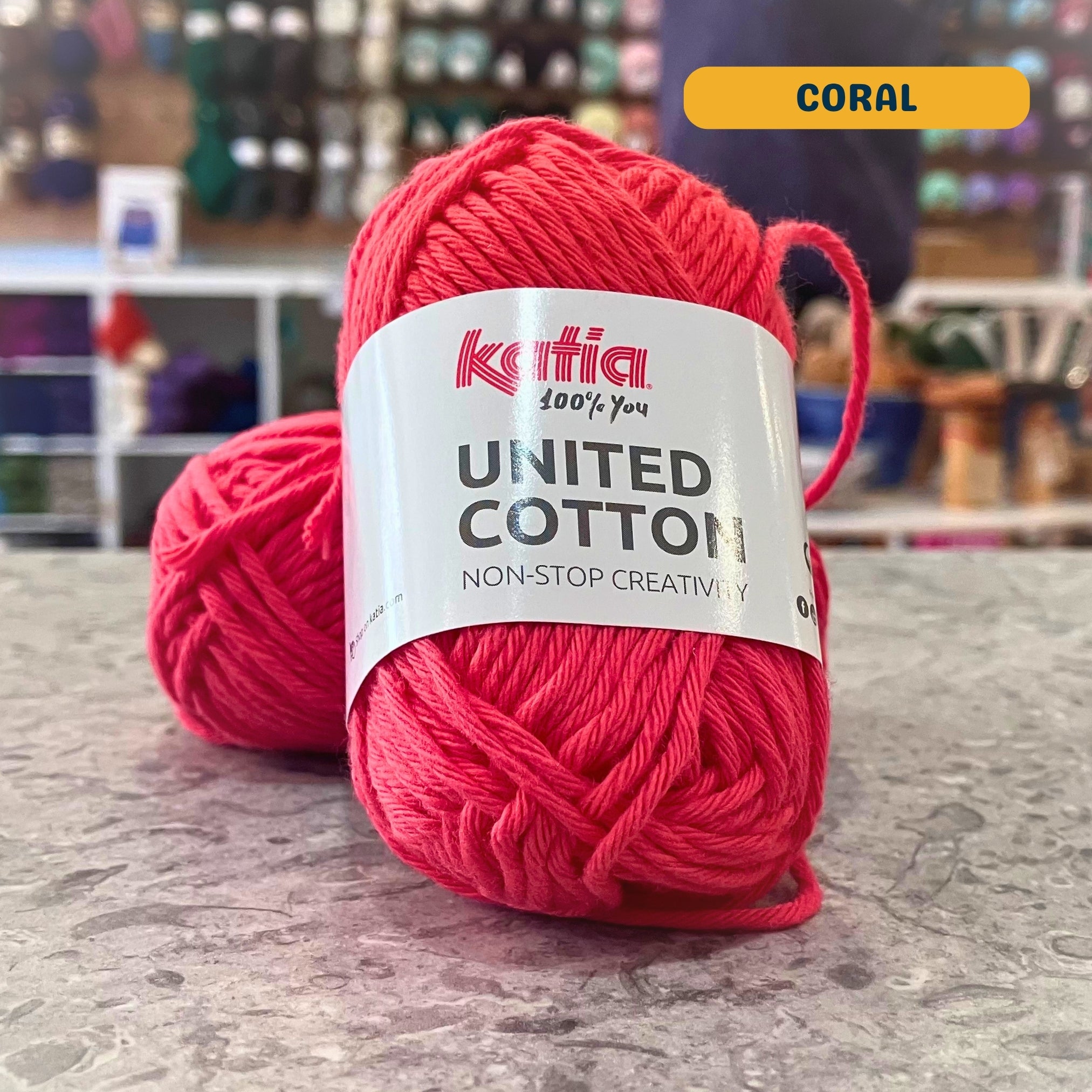 25g pink-orange (coral) cotton DK yarn, 'Katia United Cotton' label visible, craft shop background (blurred)
