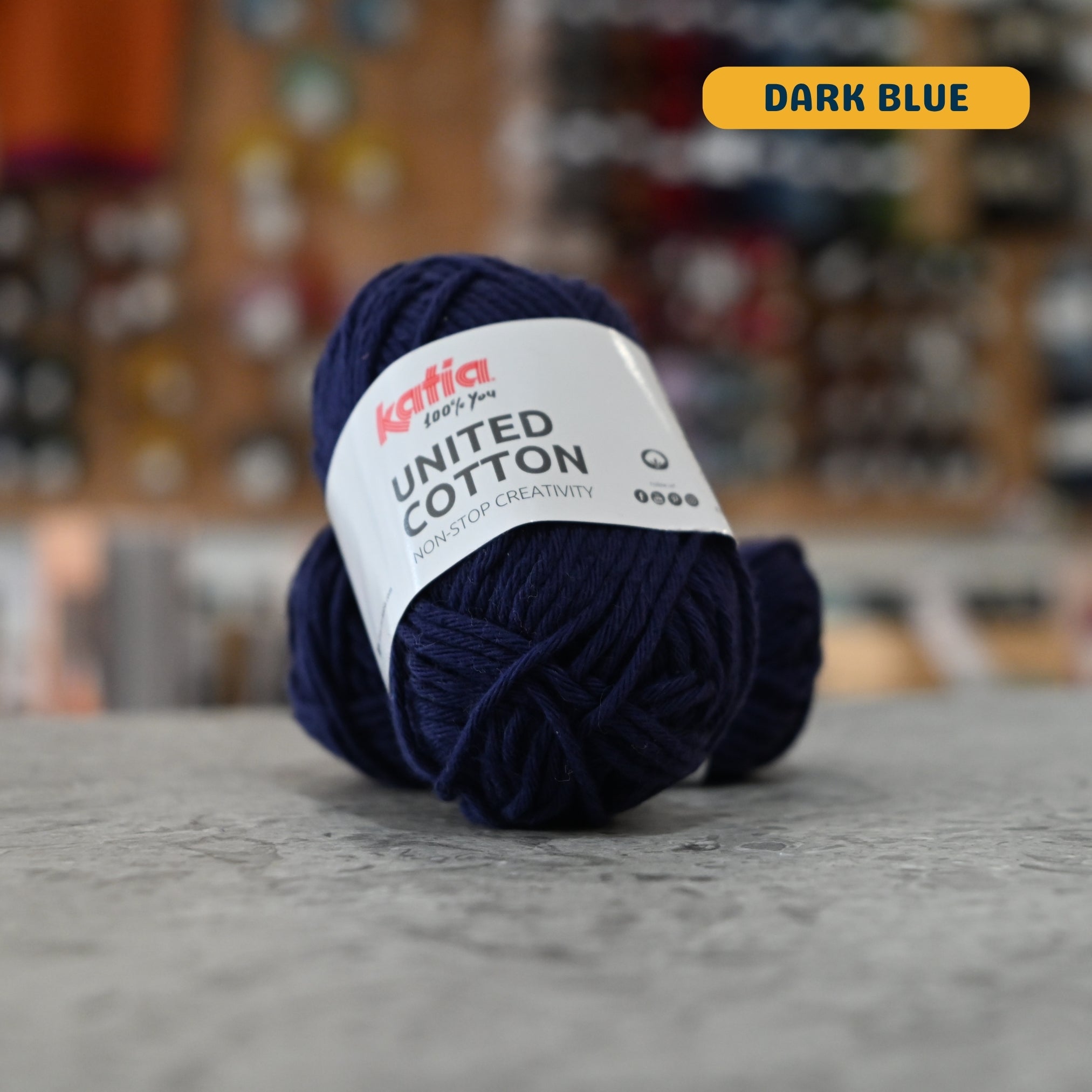 25g navy (dark blue) cotton DK yarn, 'Katia United Cotton' label visible, craft shop background (blurred)