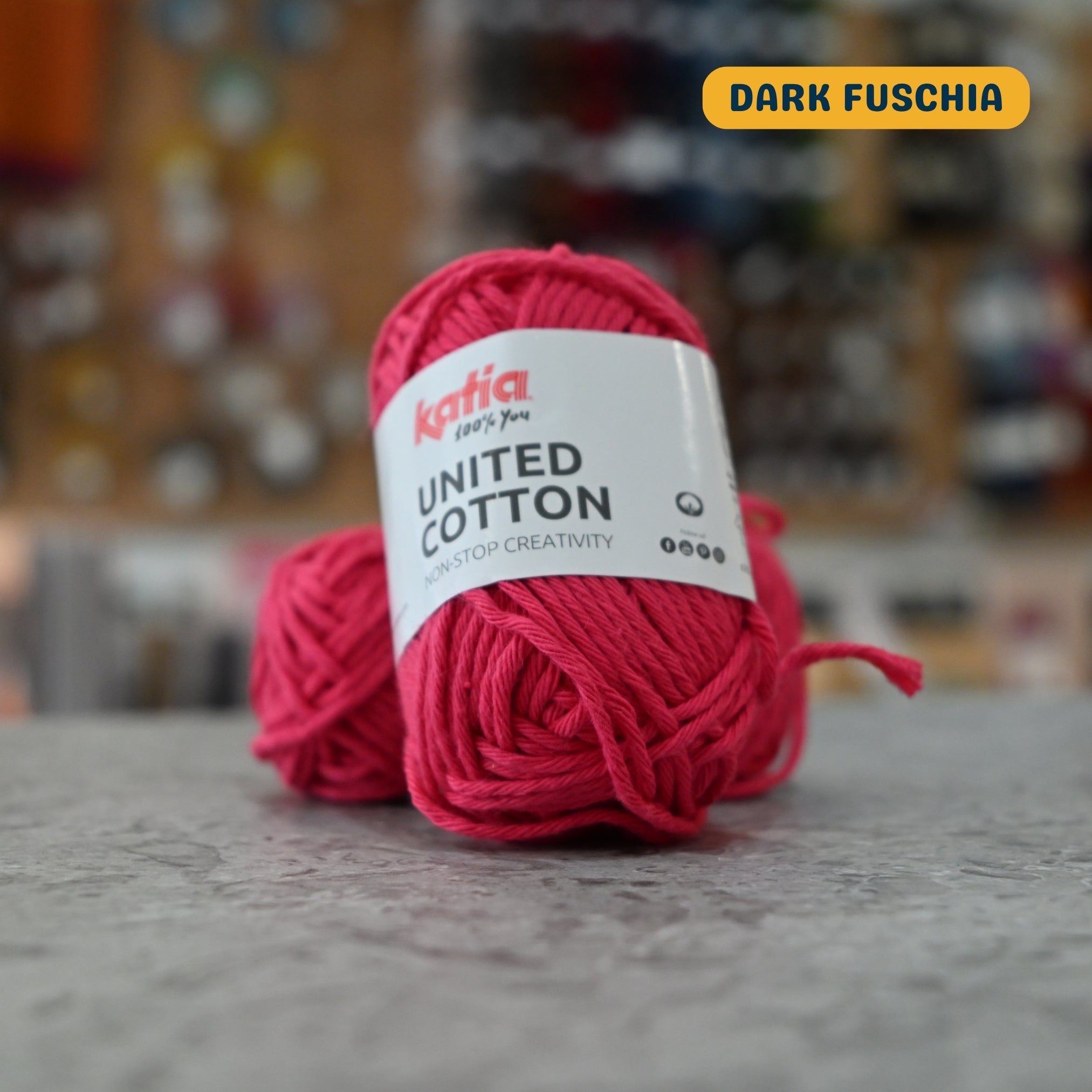 25g medium coral (dark fuschia) cotton DK yarn, 'Katia United Cotton' label visible, craft shop background (blurred)